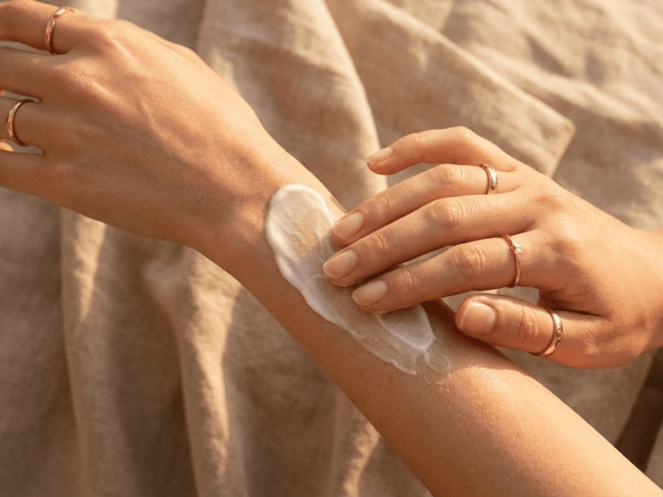 Close-up de mãos delicadas a massajar um creme hidratante rico na pele do braço, mostrando a textura suave e a absorção do produto. Iluminação solar dourada e fundo de linho bege desfocado.